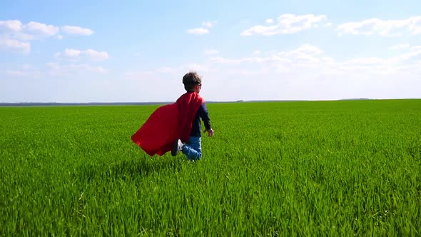 A Happy Child in a Superhero Costume in a Red Cape and in a Mask Runs on Green Grass on a Sunny Day