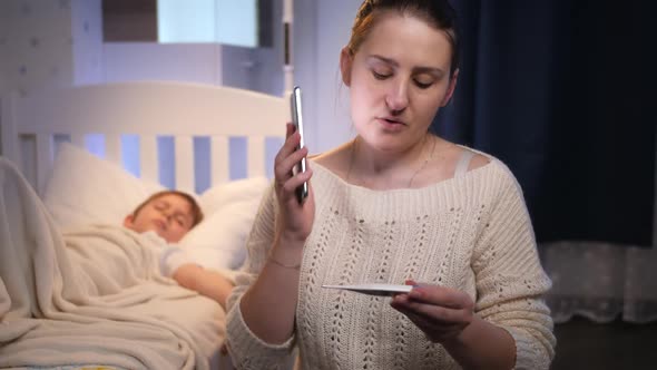 Young Woman Sitting Next to Bed of Her Sick Son Feeling Unwell and Talking to Doctor