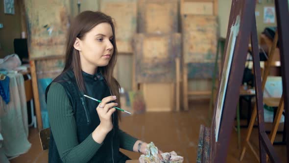 Longhaired Girl Paints with Oil Colors on Easel in Workshop Interior