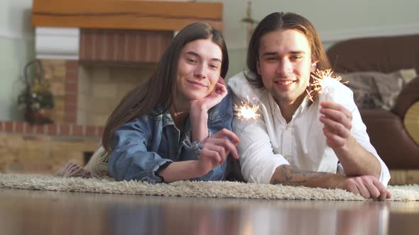 Young Beautiful Happy Guy and Girl with Sparklers in Hands Lying on the Carpet at Home