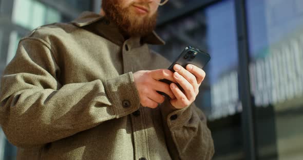 A Bearded Man is Typing on a Mobile Phone While Standing Near Office Building