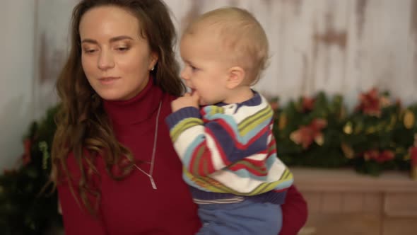 Pretty Young Woman Shakes the Baby in Her Arms Standing Near Christmas Tree and Showing Bright Toys