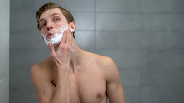 Positive Attractive Young Guy Applies Helium Foam in Front of the Mirror