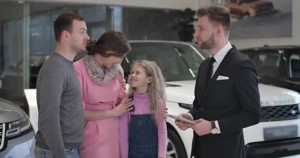 Adult Caucasian Man Talking with Trader in Car Dealership and Touching Daughter's Chin. Young Family