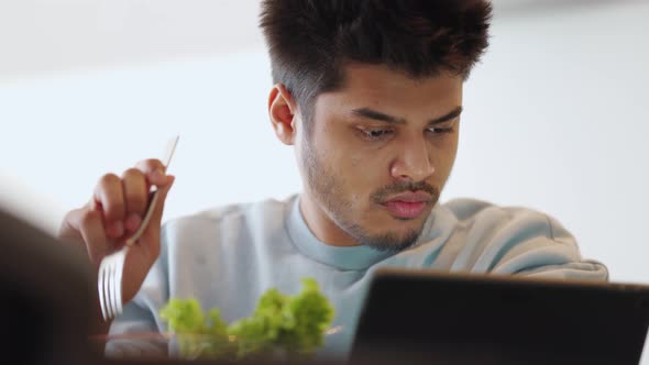 Positive Indian man wearing sweatshirt eating salad and watching something by tablet
