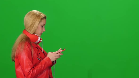 Portrait of Blonde Girl Is Walking in Big Red Headphones Texting Message on Her Mobile Phone at