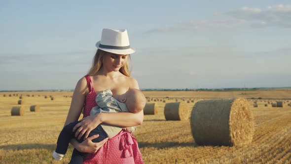 Stylish Young Mother in Hat Feeds the Baby Son with Breast in Hay Field