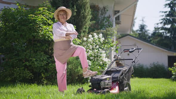 Confident Satisfied Senior Woman Crossing Hands Looking at Camera Standing with Lawn Mower Outdoors