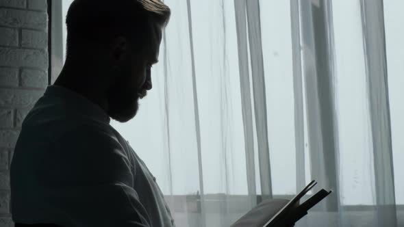 Bearded Young Man Sitting Near Window and Reading a Book at the Sunrise