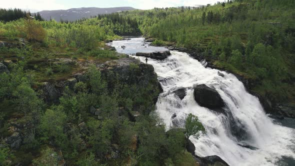 Aerial View Of Drone Operator Standing On Edge Of Cascading Waterfall Surrounded By Green Wilderness