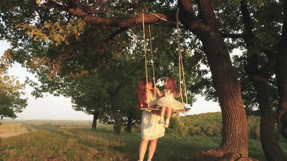 Mom Shakes Her Daughter on Swing Under a Tree in Sun. Child Laughs and Rejoices
