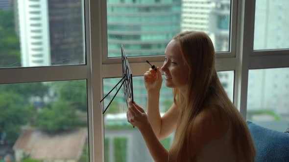 Super Slow Motion Shot of a Young Woman Doing Her Makeup Sittng By a Panoramic Window in Downtown of