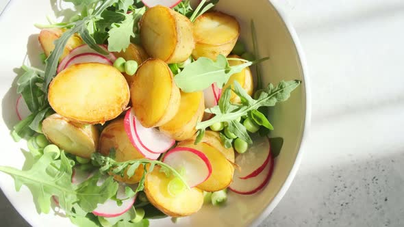 Summer potato and pea salad with arugula and radish in white bowl spinning top view.
