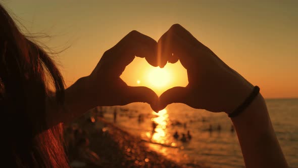 Young Beautiful Girl Makes Heart By Her Hands in Heart Shape Framing Setting Sun at Sunset Over