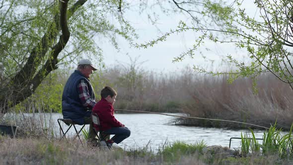 Anglers, an Elderly Man with Little Boy Catch Fish Using a Wooden Fishing Rod Sitting on Shore