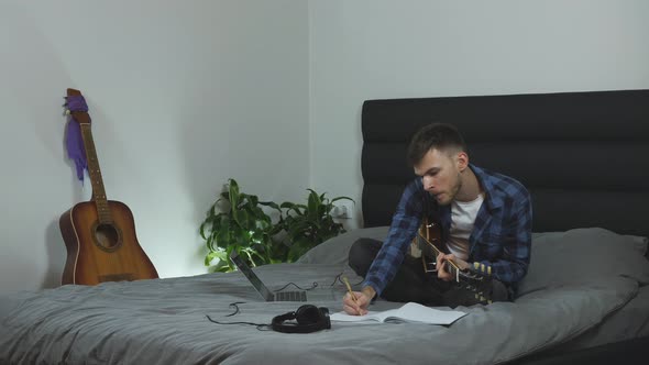 Young caucasian man in pantone blue t-shirt is learning on electric guitar on bed at modern home