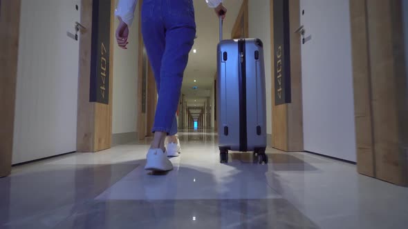 woman walks down corridor of hotel and rolls suitcase with luggage on wheels.