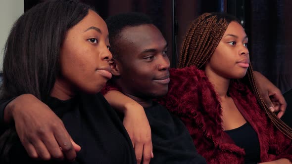 Three African Friends Watch Television in Living Room and They Hold Around Shoulders