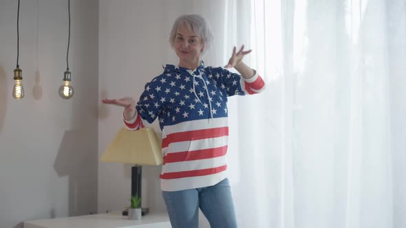 Carefree Happy Mature Woman in Casual Jeans and Stars and Stripes Hoodie Dancing in Living Room at