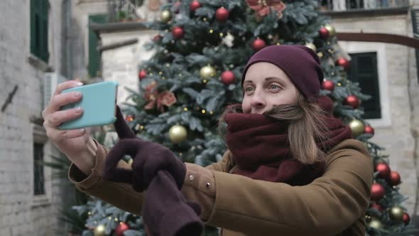 Young woman in a beige coat and a red hat photographs on the phone a Christmas tree in the old town