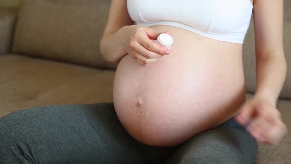 Woman Lubricates Moisturizes Pregnant Belly with Cream for Stretch Marks on Home Sofa
