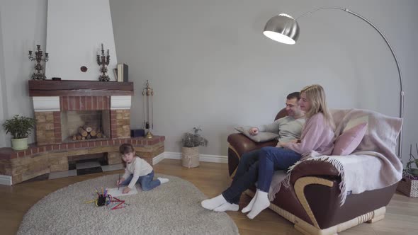 Little Brunette Caucasian Girl Sitting on Floor and Drawing As Her Parents Sitting on Armchair