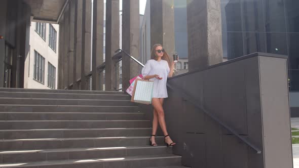 Girl with Phone Staying on Shopping Mall Stairs with Colorful Shopping Bags and Drinking Coffee