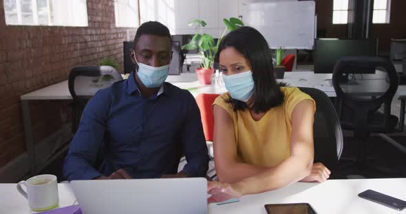 Diverse male and female business colleagues wearing face masks sitting at desk using laptop