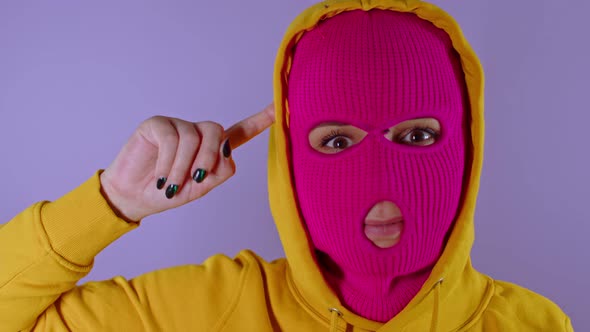 Close Up of Young Woman in Pink Balaclava and Yellow Hood Twirling Finger at Her Temple on Purple
