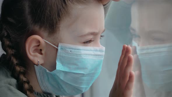 Portrait of little girl in medical mask looks sadly at her reflection in window COVID-19 infection