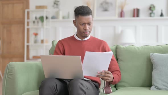 African Man with Laptop Reacting to Loss on Documents, Sofa