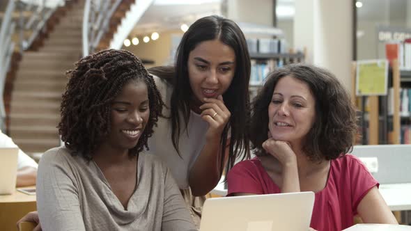 Smiling Ladies Talking While Sitting at Table with Laptop at Library