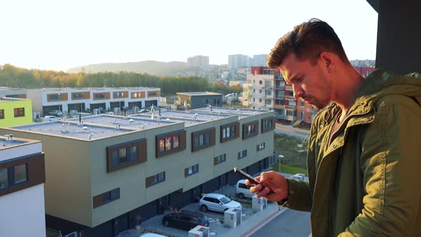 A Young, Handsome Man Types on His Smartphone on a Balcony, Eventually Looks Out