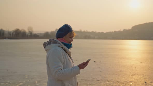 White Elderly Senior Woman Walking By the Lake with a Phone in Her Hand