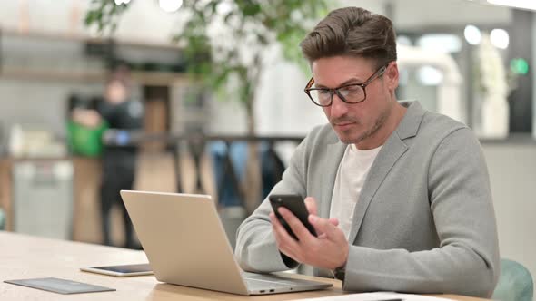 Focused Middle Aged Man with Laptop Using Smartphone 