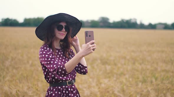Attractive Fun Hippie Woman in the Wheat Field at Sunset Making a Selfie with Smartphone