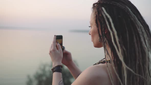 Attractive Hippie Woman with Dreadlocks Making Photo on Smart Phone at Sunset Near Sea