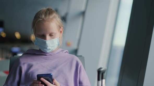 A Child in a Protective Mask Sits in the Airport Waiting Room Using a Smartphone