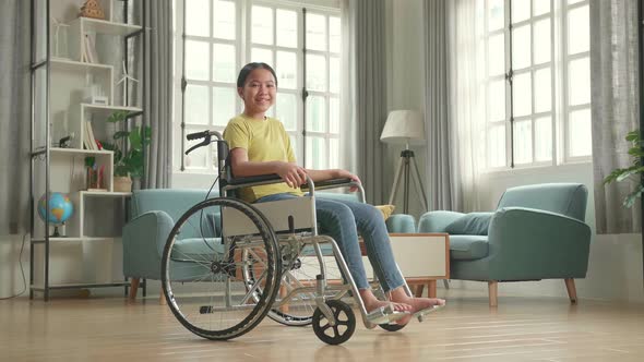 Asian Kid Girl Sitting In A Wheelchair Smiling And Looking At Camera In Living Room