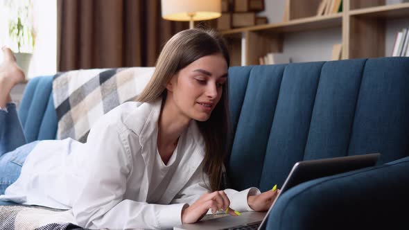 Young Woman Lying on Her Stomach a Sofa Using a Laptop Computer to Surf the Internet with a Happy