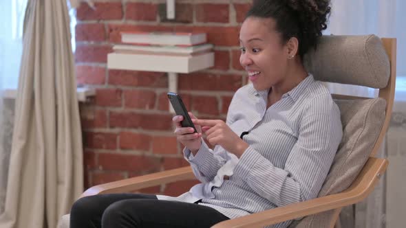 African Woman Celebrating Success on Smartphone at Home