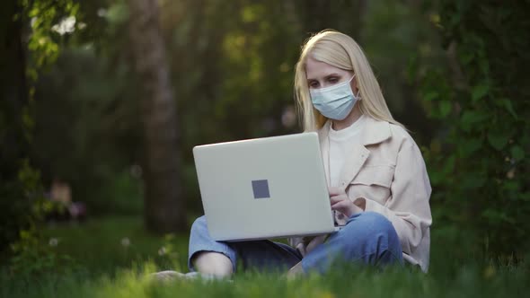 Young Woman with Curls in a Medical Mask Works Using a Laptop in the Park