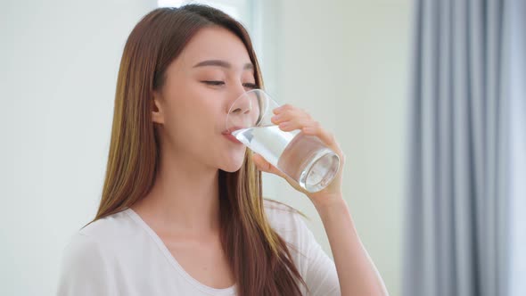 Asian attractive woman sit on bed, drink a glass of water in bedroom.