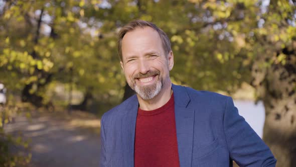 A Middleaged Handsome Caucasian Man Waves at the Camera with a Smile in a Park in Fall