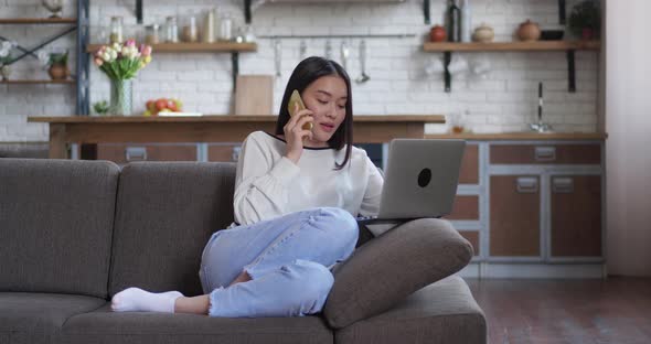 Young Asian Female Sitting in Cozy Sofa Talking on Phone at Home and Looking in Monitor of Her