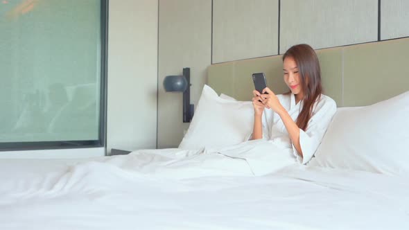 Happy Asian Woman Reading Messages on Smartphone After Waking Up in Bed. Morning Routine, Full Frame