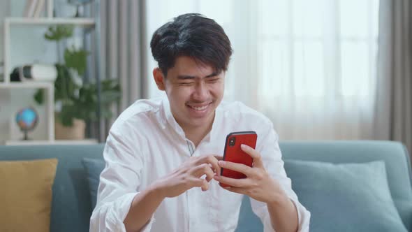 Asian Man Touching Smartphone And Laughing While Sitting On Sofa In The Living Room