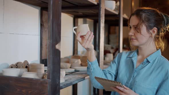 Portrait of Female Potter Checking Ceramics and Writing Information in Notebook in Workshop