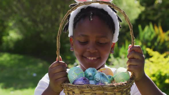 Smiling african american girl wearing easter bunny ears holding basket of easter eggs in garden