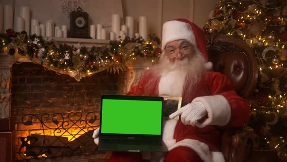 Santa Claus Holds a Laptop with Green Screen and Points at It Chroma Key While Sitting in His Rocker